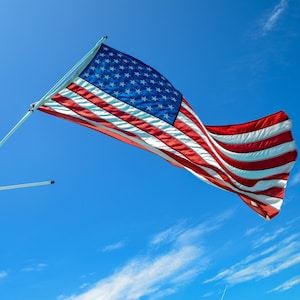 May include: An American flag waving in the wind against a bright blue sky with white clouds. The flag is attached to a white pole and is flying high above a boat.