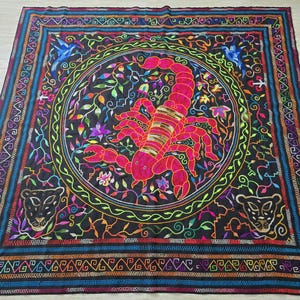 May include: A black fabric square with a colorful embroidered scorpion design in the center. The scorpion is surrounded by a circular border of flowers and vines. The fabric has a multi-colored border with geometric patterns.