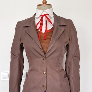 Puede incluir: Una blazer marrón con botones dorados, una camisa blanca con cuello y una corbata roja. El conjunto incluye un chaleco de punto marrón y una falda plisada azul. La ropa se exhibe en un maniquí.