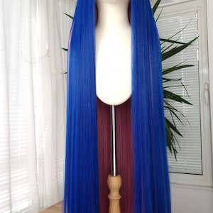 Può includere: Una lunga e vibrante parrucca blu con riflessi rosa e frangia, esposta su una testa di manichino. La parrucca ha uno stile liscio e dritto, progettata per cosplay o costumi. Il manichino è su un supporto di legno.