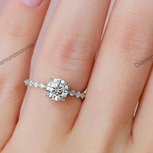 May include: A silver engagement ring with a large, round, clear gemstone. The band features small, sparkling accents. The ring is on a person's finger with light pink nail polish and daisy nail art.