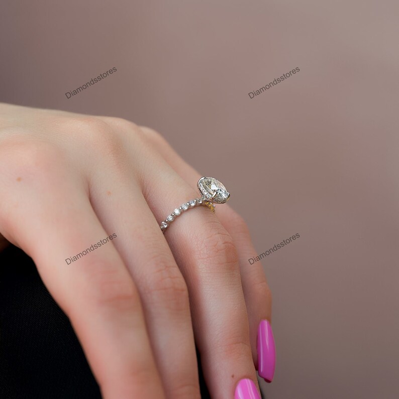 May include: A diamond engagement ring with a large oval-cut center stone and a band adorned with smaller diamonds. The ring is set in a gold and silver setting, showcasing a classic and elegant design. The ring is on a finger with pink nail polish.