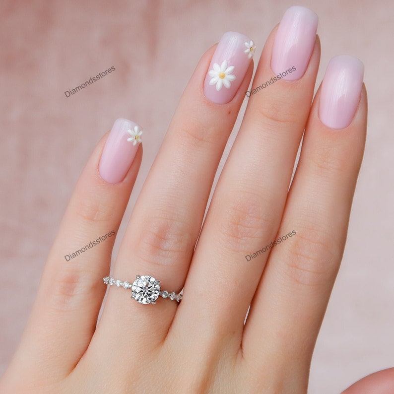 May include: A silver engagement ring with a large, round, clear gemstone. The band features small, sparkling accents. The ring is on a person's finger with light pink nail polish and daisy nail art.