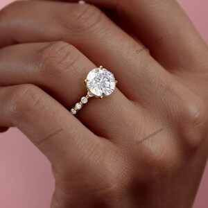 May include: A gold engagement ring featuring a large, round, brilliant-cut diamond. The ring has a delicate band with smaller diamonds set along the side. The ring is shown on a person's finger, with a soft pink background.