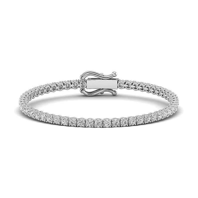 Puede incluir: Una pulsera de tenis con una fila de piedras preciosas redondas y claras engastadas en un metal plateado. La pulsera tiene un cierre de broche seguro. Las piedras preciosas brillan sobre un fondo blanco.