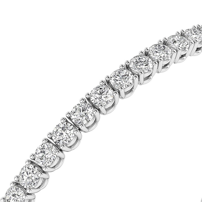 Puede incluir: Primer plano de una pulsera de tenis con una serie de diamantes redondos con talla brillante engastados en metal blanco. Los diamantes brillan sobre un fondo blanco, mostrando el elegante dise&ntilde;o de la joya.