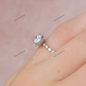 May include: A silver engagement ring with a large, oval-cut diamond center stone. The ring features a halo of smaller diamonds around the center stone and a delicate band with graduated diamonds. The ring is set on a finger.