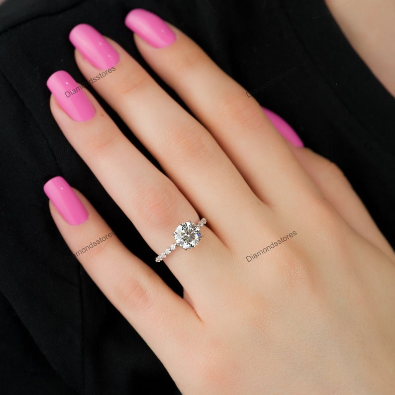 May include: A silver ring with a large, round, clear gemstone as the centerpiece. The band is adorned with smaller, clear gemstones. The ring is on a hand with pink nail polish, set against a black background.