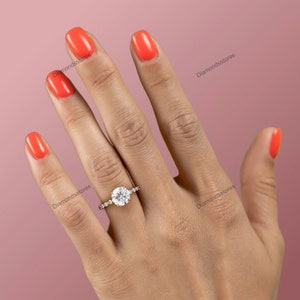 May include: A gold ring with a large, round, clear gemstone as the centerpiece. The ring is set with smaller, clear gemstones on either side. The background is a solid pink color.