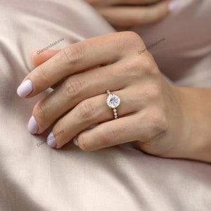 May include: A gold engagement ring featuring a large, round, clear gemstone as the centerpiece. The band is adorned with smaller, clear gemstones. The ring is on a person's finger, set against a soft, neutral background.