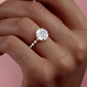 May include: A rose gold engagement ring with a large, round, clear gemstone as the centerpiece. The band features smaller, round gemstones set in a delicate row. The ring is shown on a person's finger against a soft pink background.