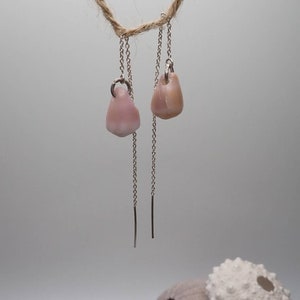 Puede incluir: Un par de delicados pendientes de cadena de plata con colgantes de concha rosa. Las conchas son de color rosa claro y tienen una forma natural y orgánica.