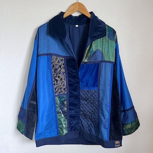 Puede incluir: Una chaqueta patchwork en tonos azules y verdes cuelga de una percha de madera. La chaqueta presenta un cuello de terciopelo y paneles de varias texturas y patrones, incluyendo telas acolchadas y estampadas. El diseño general es una mezcla de texturas y colores.