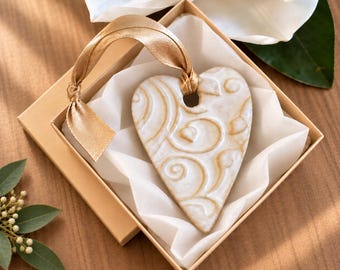 May include: A heart-shaped ceramic ornament featuring a glossy white finish with embossed gold-toned swirl patterns. The ornament is attached to a metallic gold ribbon and is presented inside a square gift box lined with white tissue paper.