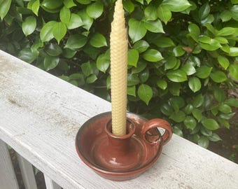 May include: A tall, light yellow, tapered beeswax candle with a textured honeycomb pattern sits upright in a round, glossy, reddish-brown ceramic candle holder. The holder features a small, curved handle on one side and a central socket.