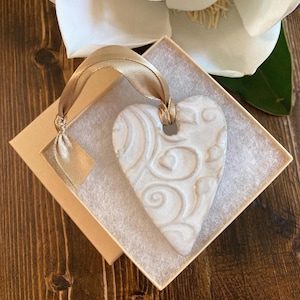 May include: A white ceramic heart ornament featuring an embossed swirling pattern on its surface. The heart is suspended by a metallic gold ribbon loop and is presented inside a square cardboard gift box lined with white protective padding.