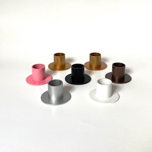 May include: A collection of seven cylindrical candle holders with flat circular bases, arranged in a group. The holders are finished in various solid colors including pink, gold, black, silver, white, and brown, each featuring a smooth surface.
