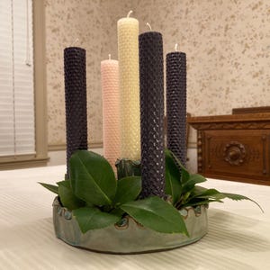 May include: A circular, light green ceramic candle holder containing five tall, cylindrical beeswax candles with a textured honeycomb pattern. The candles are arranged in a cluster, featuring two dark purple, one light pink, one cream, and one yellow.