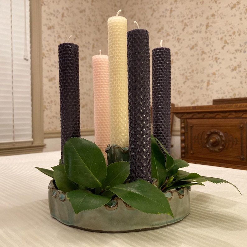 May include: A circular, light green ceramic candle holder containing five tall, cylindrical beeswax candles with a textured honeycomb pattern. The candles are arranged in a cluster, featuring two dark purple, one light pink, one cream, and one yellow.