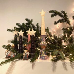 May include: A collection of honeycomb-textured beeswax candles shaped like Christmas trees, topped with stars in cream and pink. One candle sits in a hand-painted ceramic candlestick with floral motifs, accompanied by three small figurines.