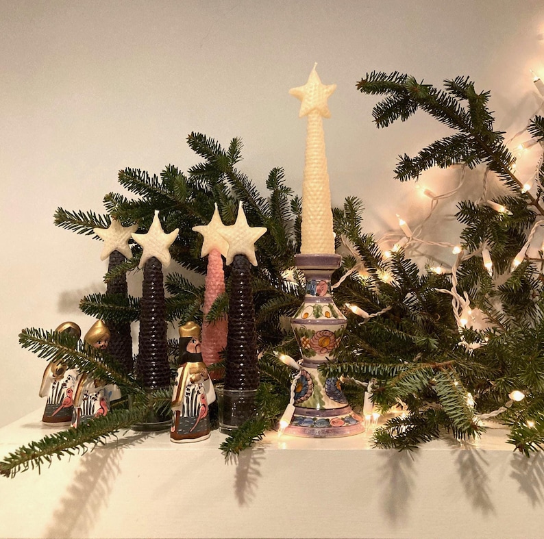May include: A collection of honeycomb-textured beeswax candles shaped like Christmas trees, topped with stars in cream and pink. One candle sits in a hand-painted ceramic candlestick with floral motifs, accompanied by three small figurines.