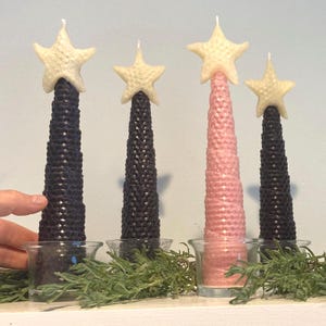 May include: Four conical candles with a honeycomb texture, three in black and one in light pink, each topped with a cream-colored star. The candles are placed in clear glass votive holders, with green sprigs arranged around the base of each candle.