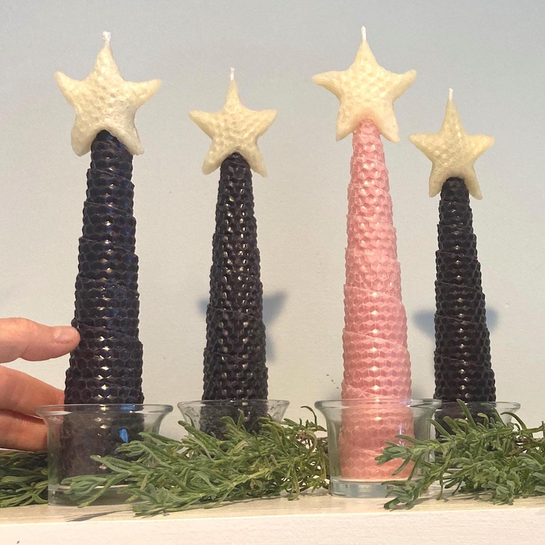 May include: Four conical candles with a honeycomb texture, three in black and one in light pink, each topped with a cream-colored star. The candles are placed in clear glass votive holders, with green sprigs arranged around the base of each candle.