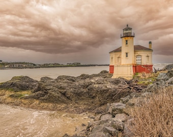 Pode incluir: Um farol branco com uma base vermelha fica em um afloramento rochoso. O farol é cercado por água e o céu está nublado com nuvens escuras.