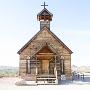 May include: A wooden church with a steeple and a cross on top. The church is made of weathered wood and has a porch with a wooden railing. The church is surrounded by a desert landscape.