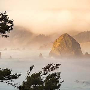 May include: A large rock formation, known as Haystack Rock, rises from the ocean, partially obscured by fog. The rock is surrounded by smaller rock formations and the ocean is a light blue color. The sky is a pale pink and orange color, with clouds.