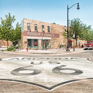 May include: A red truck is parked on a street corner in front of a building with the words "Winslow Arizona" on the side. The street is marked with a large painted Route 66 sign.