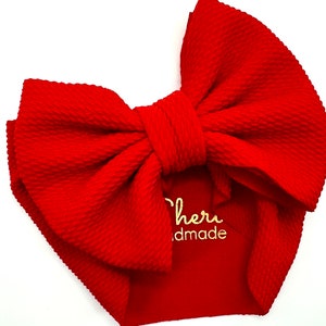 Peut inclure: Un bandeau de bébé rouge vif avec un grand nœud. Le bandeau présente un motif texturé en forme de gaufre. Le texte "Cheri handmade" est visible sur le tissu.