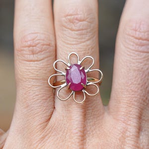 Può includere: Anello dorato con una grande gemma di rubino taglio ovale. Il rubino è incastonato in un delicato design floreale traforato. L'anello è indossato su un dito, mettendo in risalto il design del gioiello.