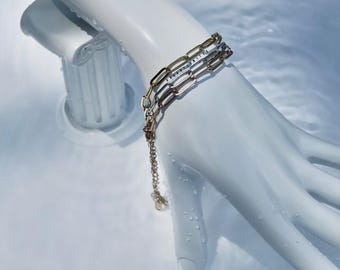 May include: A gold and silver chain bracelet with a delicate chain and a small charm. The bracelet is displayed on a white hand, with a white background.