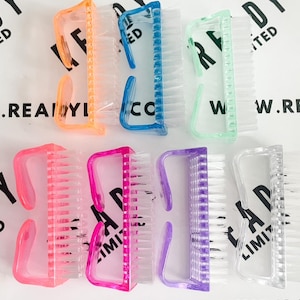 May include: A set of ten small, clear plastic brushes with colourful handles. The brushes have a rectangular shape and are designed for cleaning and scrubbing. The handles are in various colours, including orange, blue, green, pink, and purple. The brushes are arranged in two rows, with the handles facing up.