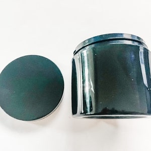 May include: A black, round, lid for a jar or container. The lid is smooth and glossy.