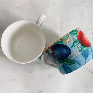 May include: Two white ceramic mugs. One mug is plain white, the other has a blue and green design with red and blue fruit.