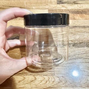 Puede incluir: Un tarro de plástico transparente y cilíndrico con tapa negra. El tarro está sostenido por una mano y está vacío. El tarro está diseñado para almacenamiento y organización.