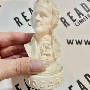 May include: A small, off-white bust of Mozart. The bust features a detailed depiction of Mozart's face, hair, and clothing. The base of the bust is inscribed with the name "MOZART". The bust is held in a hand.