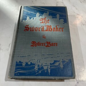 May include: A vintage book titled "The SwordMaker" by Robert Barr. The book has a blue and grey cover with red lettering. The cover features a blue silhouette design of a city skyline.