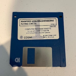 May include: A blue 3.5 inch floppy disk labeled "Manfred von Krashenberns Flying Circus" with the text "Disk 1 IBM-585 Computer Program Diskette For use with IBM PC/TANDY® and Compatible Computer Systems COSMI 1994 COSMI CORP MFG. IN U.S.A."