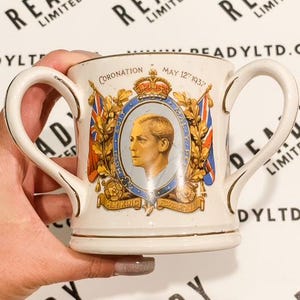 May include: A white ceramic mug with a gold rim and two handles. The mug features a commemorative design for the coronation of King Edward VIII on May 12, 1937. The design includes a portrait of the King, the royal coat of arms, and the text "Coronation - May 12th 1937".