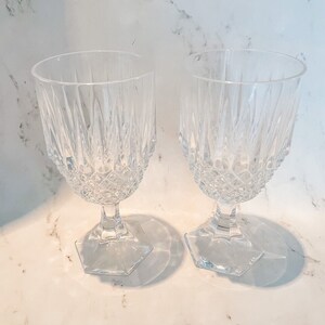 May include: Two clear crystal wine glasses with a diamond and vertical line pattern. The glasses have a hexagonal base and a stem. The glasses are empty and sit on a white marble surface.