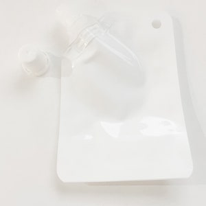 Puede incluir: Una bolsa de plástico blanca con un pico de plástico transparente y una tapa blanca. La bolsa tiene un agujero en la parte superior para colgarla.