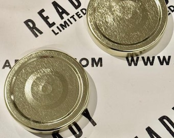 May include: Two gold metal jar lids with a textured surface. The lids have a raised circle in the center. The lids are on a white surface with the text "READY LIMITED" printed on the surface.