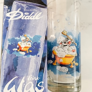May include: A clear glass drinking glass with a blue and white cartoon illustration of a dog in a bathtub. The glass is packaged in a blue box with the text "Thomas Gottschalk Diddl Trink-Glas" printed on it.