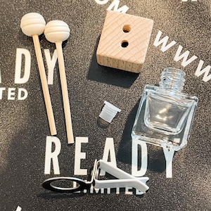 Puede incluir: Un conjunto de suministros para ambientador de coche que incluye una botella de vidrio transparente, un bloque difusor de madera con dos agujeros, dos palitos de madera, una pinza de metal y una pequeña tapa de plástico blanco. Los artículos están dispuestos sobre una superficie negra con el texto "READY LIMITED" y "www.com" impreso en ella.