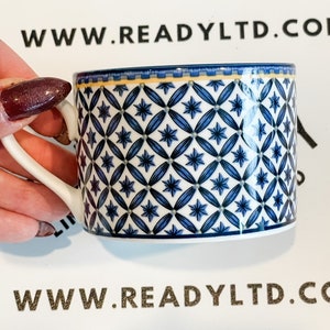 Pode incluir: Uma chávena de café de cerâmica branca com um padrão geométrico azul e branco. A chávena tem uma borda dourada e uma banda dourada em torno da base. A chávena está sobre uma superfície branca com o texto "www.readyltd.co" impresso em dourado.