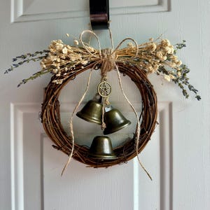May include: A rustic grapevine wreath adorned with dried flowers, lavender sprigs, and three bronze-colored bells. A jute bow and a pentacle charm add decorative detail. The wreath hangs on a white door.