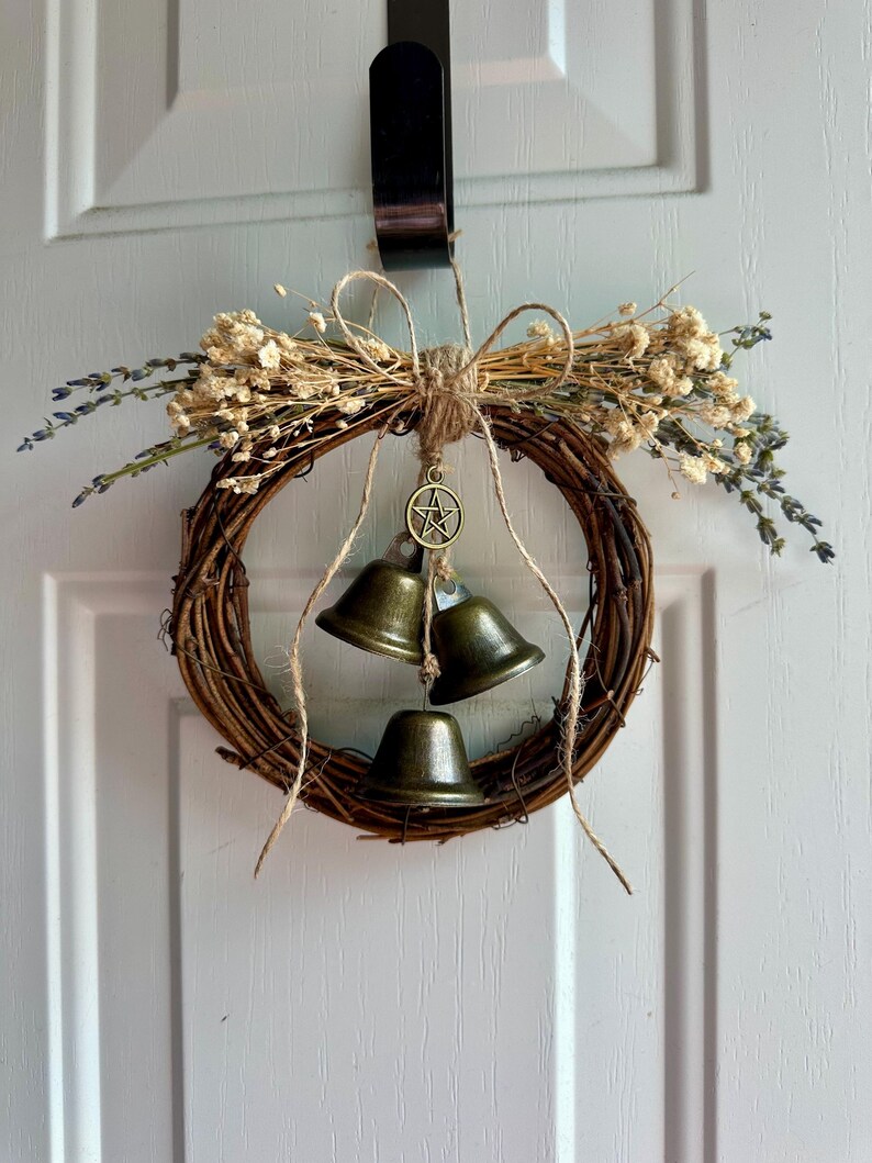 May include: A rustic grapevine wreath adorned with dried flowers, lavender sprigs, and three bronze-colored bells. A jute bow and a pentacle charm add decorative detail. The wreath hangs on a white door.